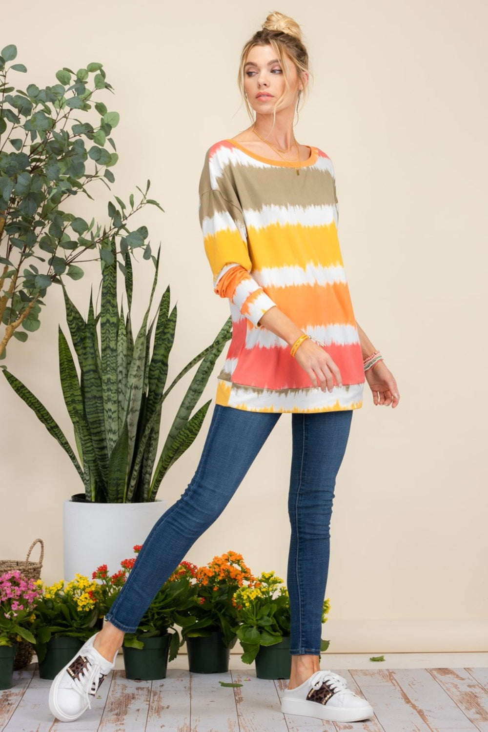 Wearing the Celeste Full Size Striped Long Sleeve T-Shirt paired with jeans, a person smiles while standing against a light background adorned with plants, exuding a casual yet stylish look.