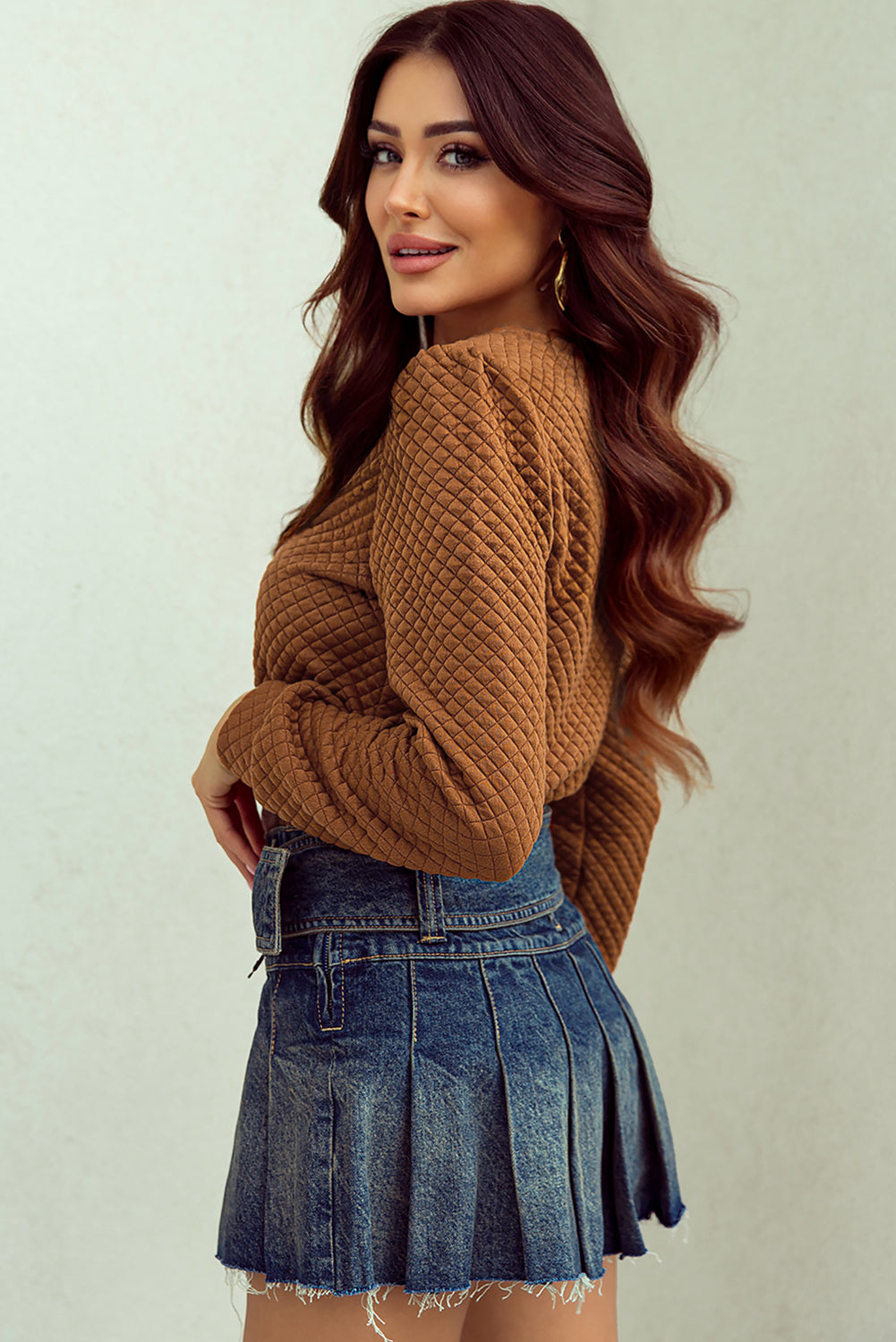 A woman with long, wavy brown hair stands in front of a light-colored background, looking over her shoulder and smiling, while wearing a Brown Solid Color Quilted Puff Sleeve Pullover Sweatshirt and a pleated denim skirt.