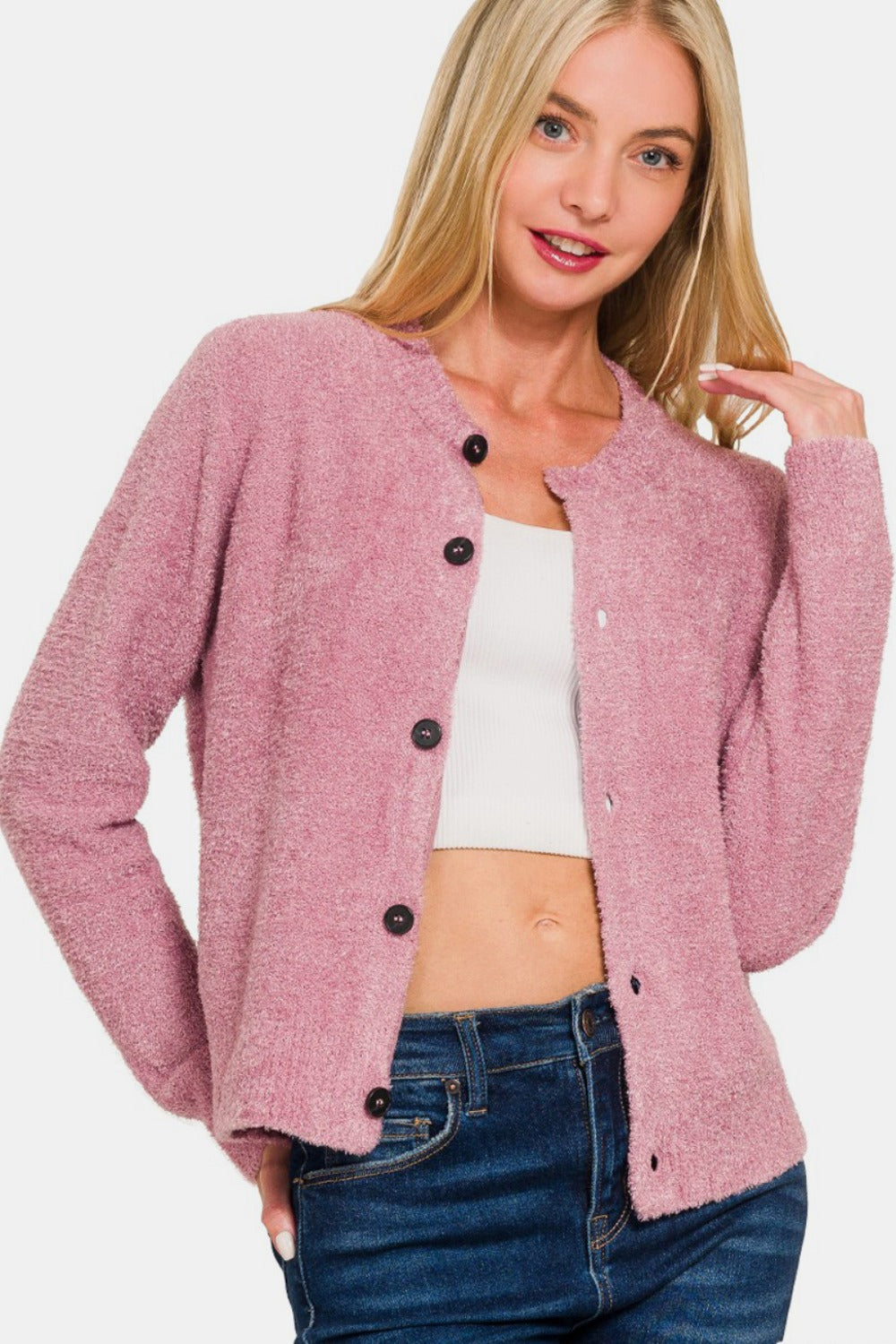 A person with long blonde hair is wearing a Zenana Button Down Long Sleeve Sweater Cardigan in pink over a white crop top and blue jeans, posing with one hand touching their hair.