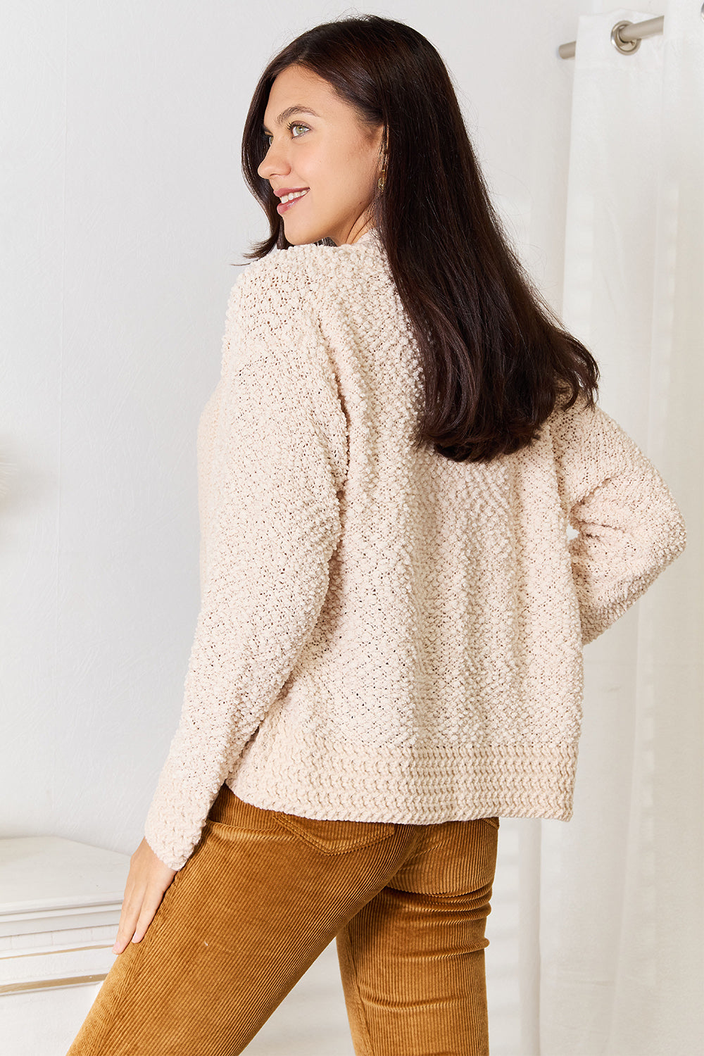 A person with long braided hair is standing and wearing the Double Take Open Front Long Sleeve Cardigan in beige over a white top, paired with mustard-colored pants. They are looking to the side, holding the comfortable fabric of the cardigan together.