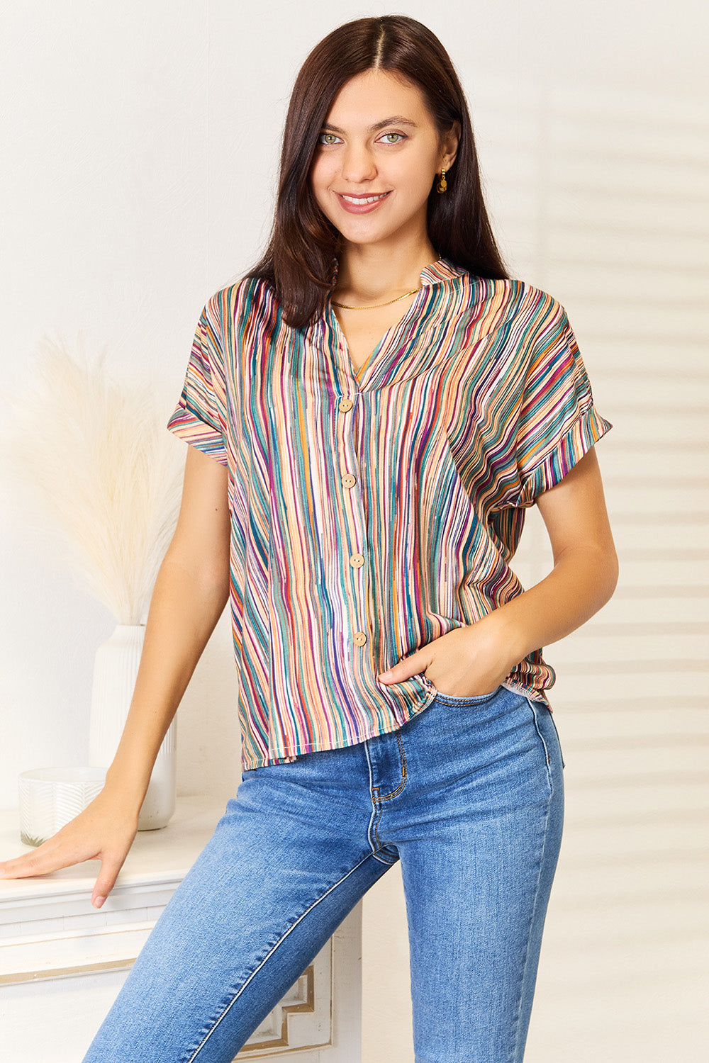 A person with long, curly hair wearing a Shiny Multicolored Stripe Notched Neck Top and blue jeans stands with one hand in their pocket, smiling slightly.