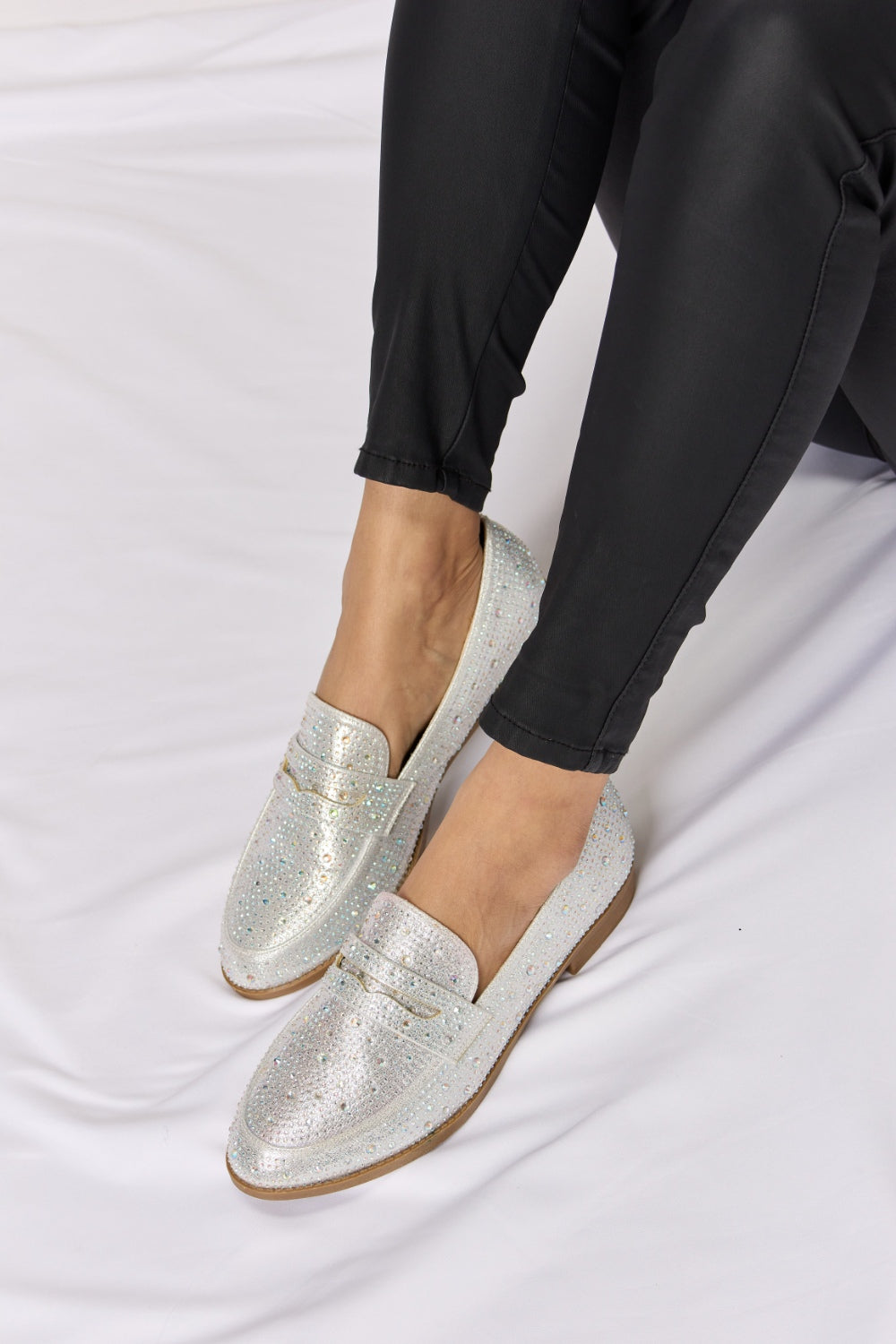 A person wearing black pants and elegant Forever Link Rhinestone Point Toe Loafers sits gracefully on a white surface.