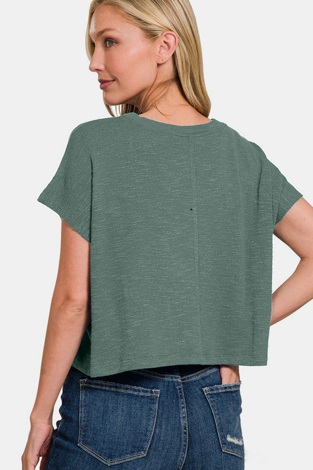 A woman with long blonde hair is wearing a green, short-sleeve Zenana Round Neck Crop T-Shirt and blue jeans. This versatile wardrobe staple complements her ensemble perfectly. She stands with a neutral expression against a plain white background.