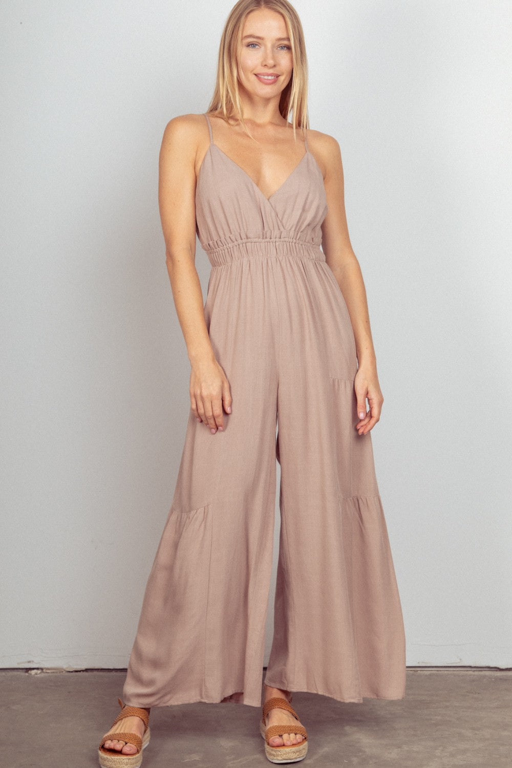 A woman standing indoors wearing the VERY J Sleeveless Ruched Wide Leg Jumpsuit in beige with adjustable straps and sandals, set against a light gray background.