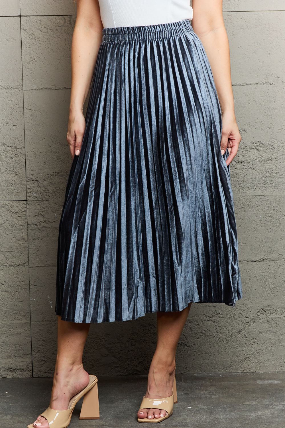 A person in a white top and the Ninexis Accordion Pleated Flowy Midi Skirt stands against a textured wall, wearing beige heeled sandals.