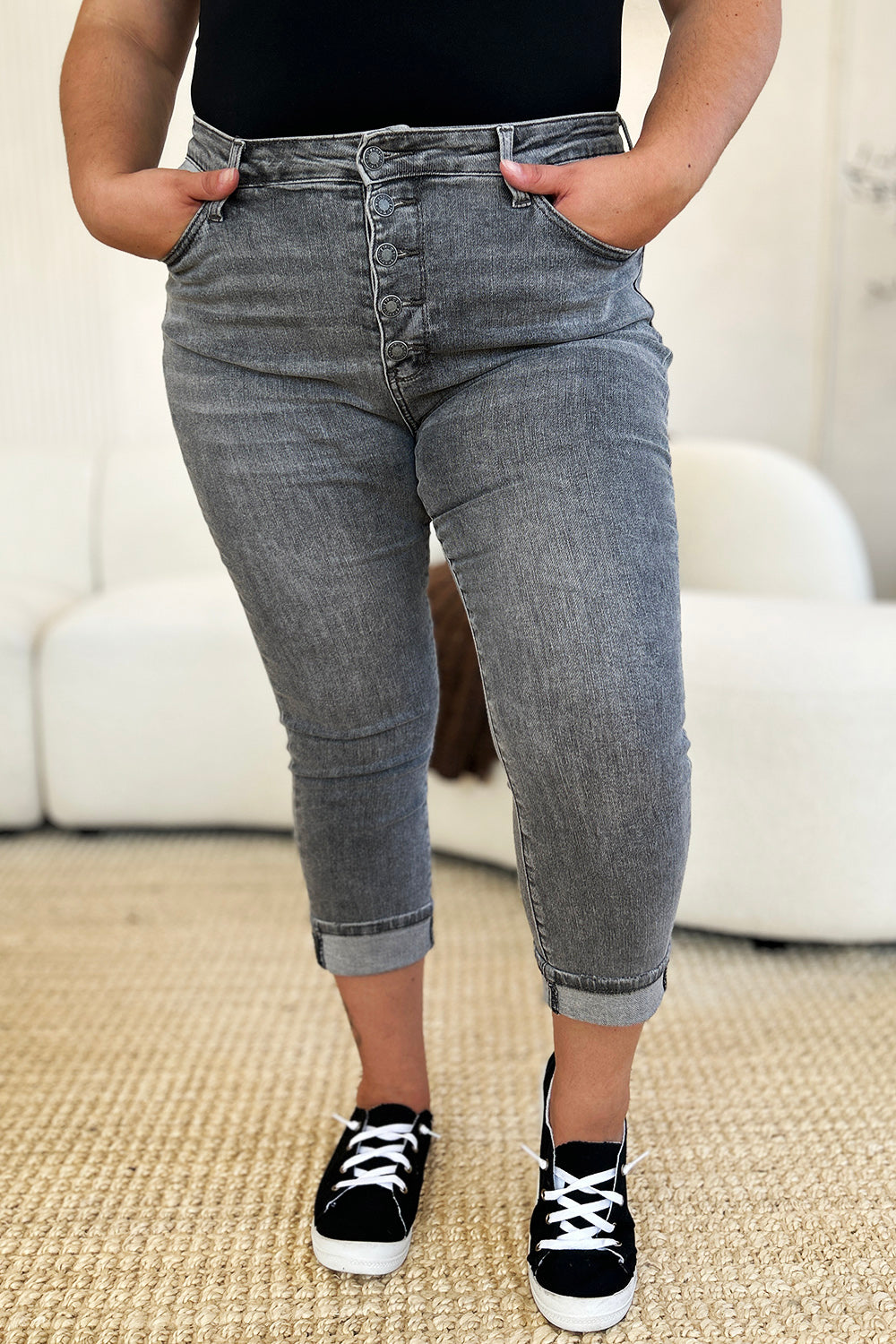 A person wearing Judy Blue Full Size Button Fly High Waist Cuffed Capris in gray, paired with a white top and white sneakers, stands on a beige carpeted floor.