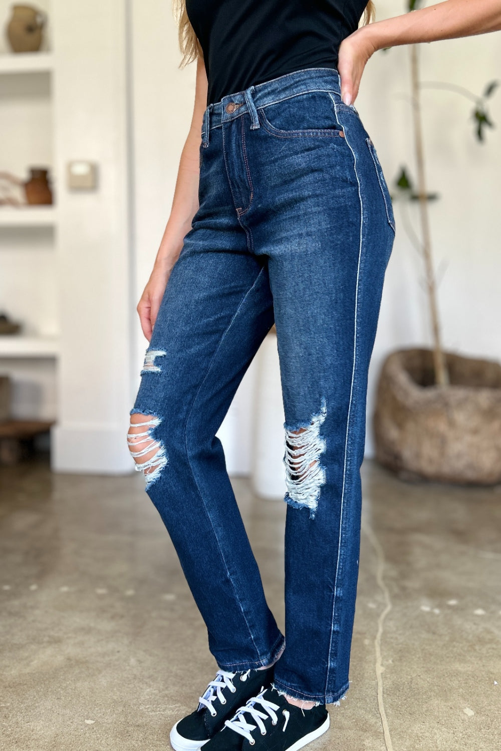 A woman in a black sleeveless top and Judy Blue Full Size High Waist Rigid Magic Heavy Destroy Straight Jeans stands indoors with her hands in her pockets. The background features a plant and shelves adorned with decor items.