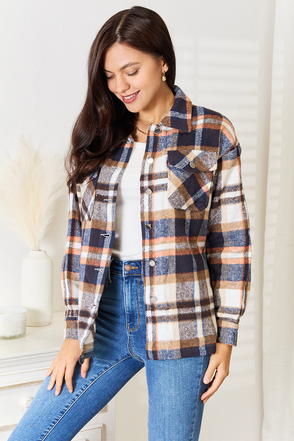 A person with long braided hair wearing the Double Take Plaid Button Front Shirt Jacket with Breast Pockets over a white top and blue jeans stands with a hand in their pocket, posing in an indoor setting. The layering of the jacket adds an effortless touch to the outfit.