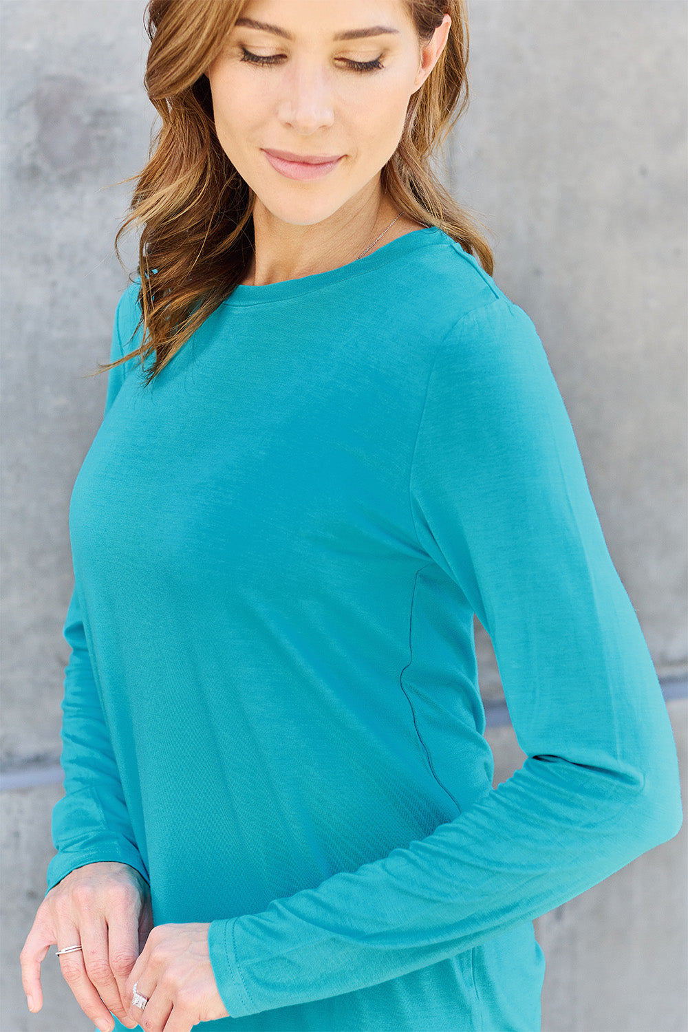 A woman with shoulder-length hair wearing the Basic Bae Full Size Round Neck Long Sleeve Top and blue jeans stands in front of a concrete wall, holding a beige handbag over her shoulder. Her outfit exemplifies basic style, with the top made from 100% polyester and recommended for machine wash cold.