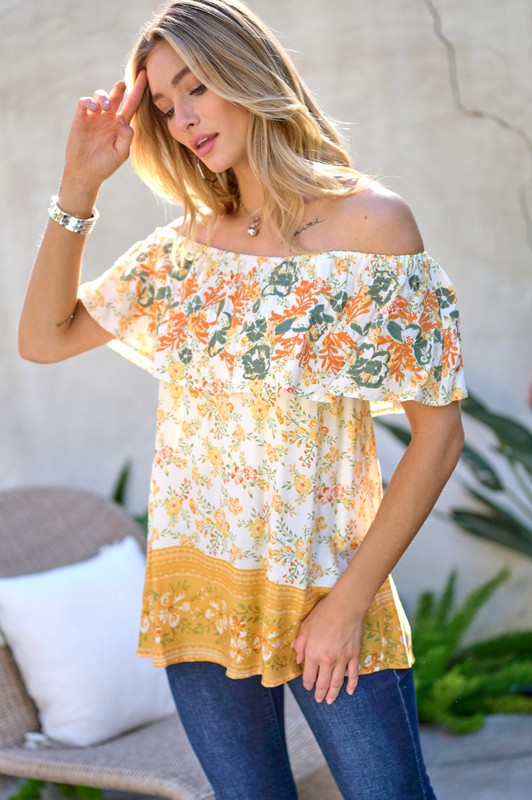 A woman with blonde hair is smiling while standing outdoors, wearing the Printed Off Shoulder Smocked Top paired with jeans.