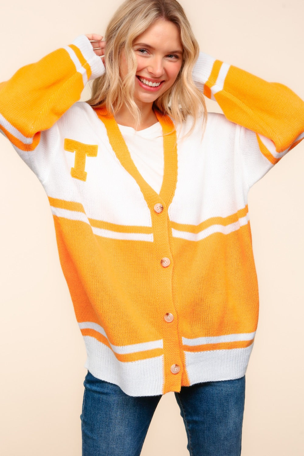 A person wearing the Haptics V Neck Button Down Letter Patch Cardigan, oversized with white and orange stripes and a yellow "T" patch, paired with blue jeans, smiles while lifting their hands to their head.