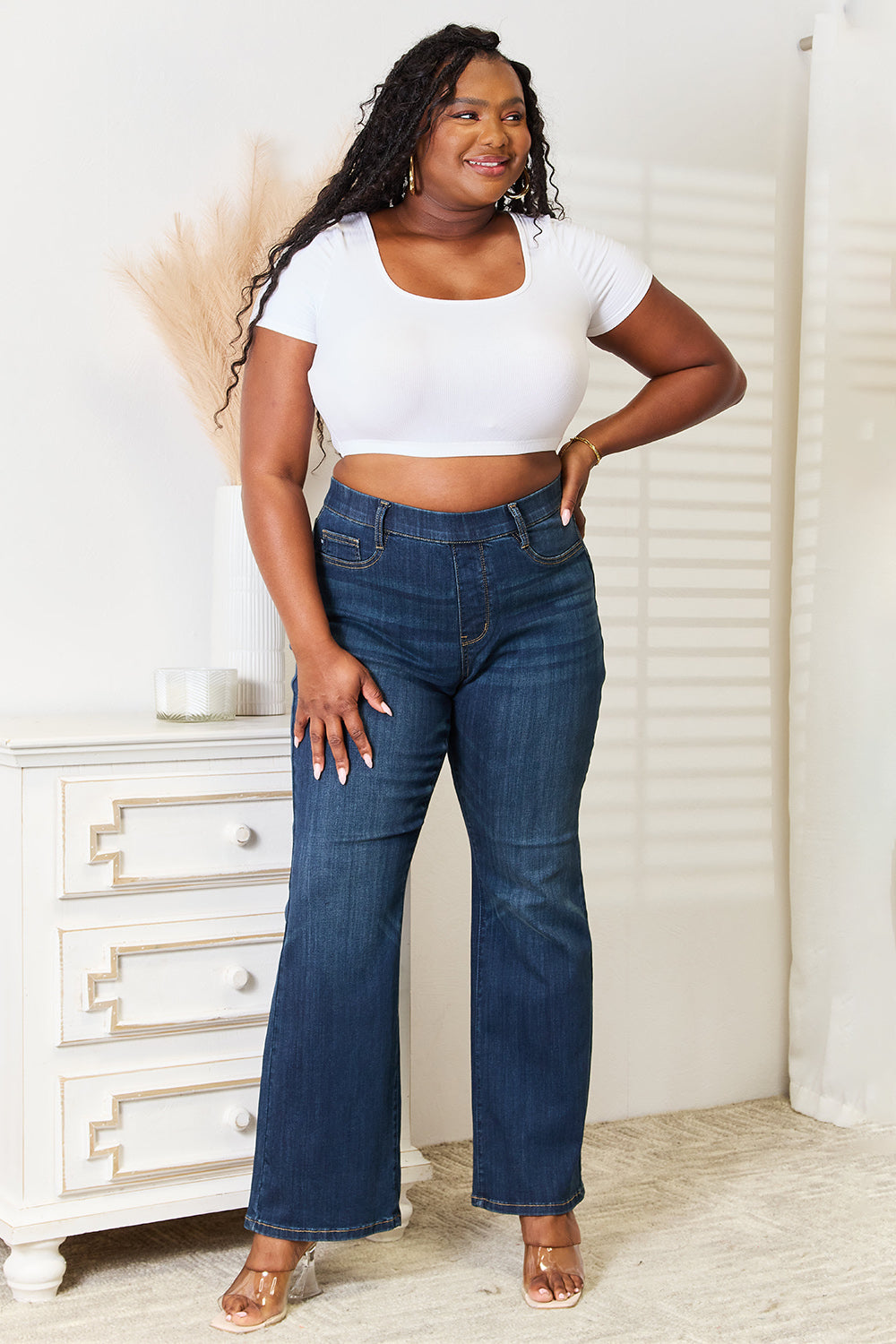 A person wearing a white crop top and Judy Blue Full Size Elastic Waistband Slim Bootcut Jeans stands next to a white dresser.