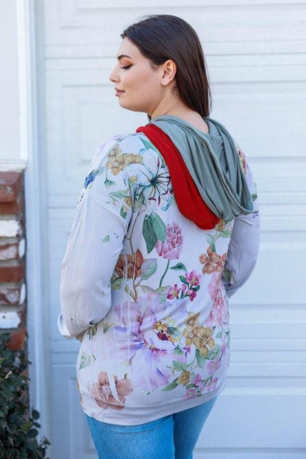 A woman stands outdoors wearing the Blumin Apparel Full Size Floral Zip-Up Hoodie with her hands in the pockets. A leafy green hedge is in the background, adding to the casual chic vibe.