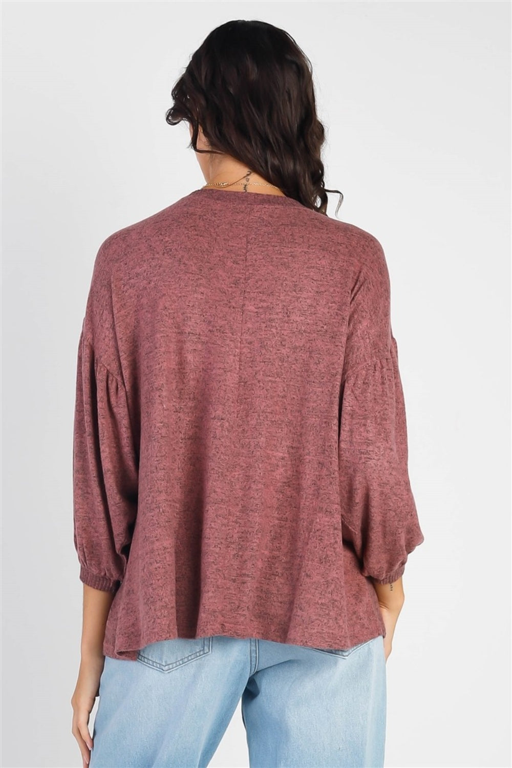 A person with long dark hair stands against a white background, smiling and raising one hand while wearing the trendy and stylish Cherish Apparel Drop Shoulder Puff Sleeve Top in burgundy paired with light blue jeans.