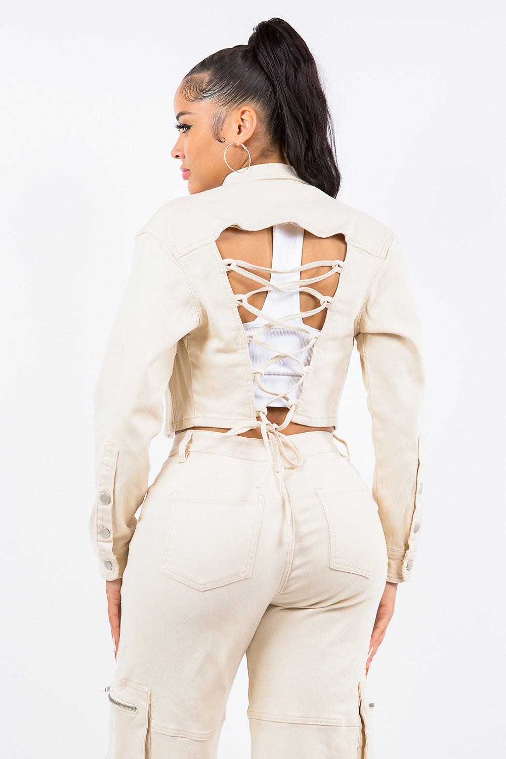 A person wearing an American Bazi Laced Back Cropped Jacket in beige with matching pants and a white crop top stands against a plain white background. They have long dark hair tied in a high ponytail and are looking towards the camera.