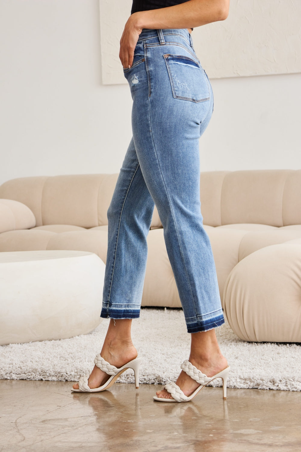 A person modeling Judy Blue Full Size Release Hem Cropped Bootcut Jeans and white heeled sandals stands in a room with light-colored furniture.