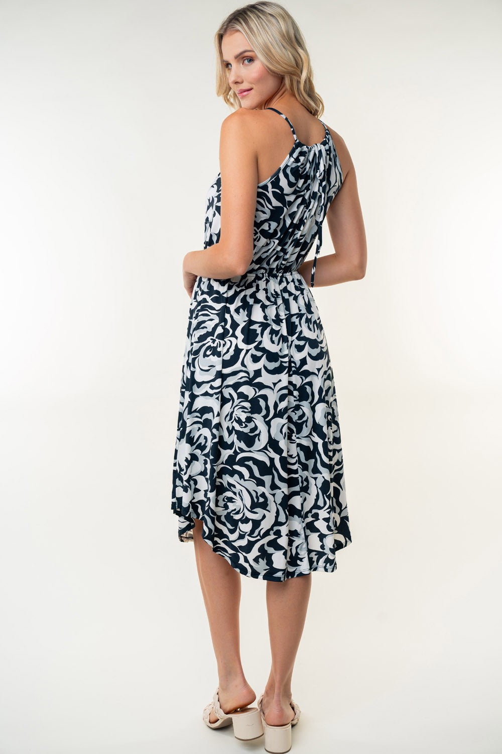 The person wearing a White Birch Tied Ruched Floral Sleeveless Knee Length Dress, featuring a black and white floral pattern, stands and smiles against a neutral background.