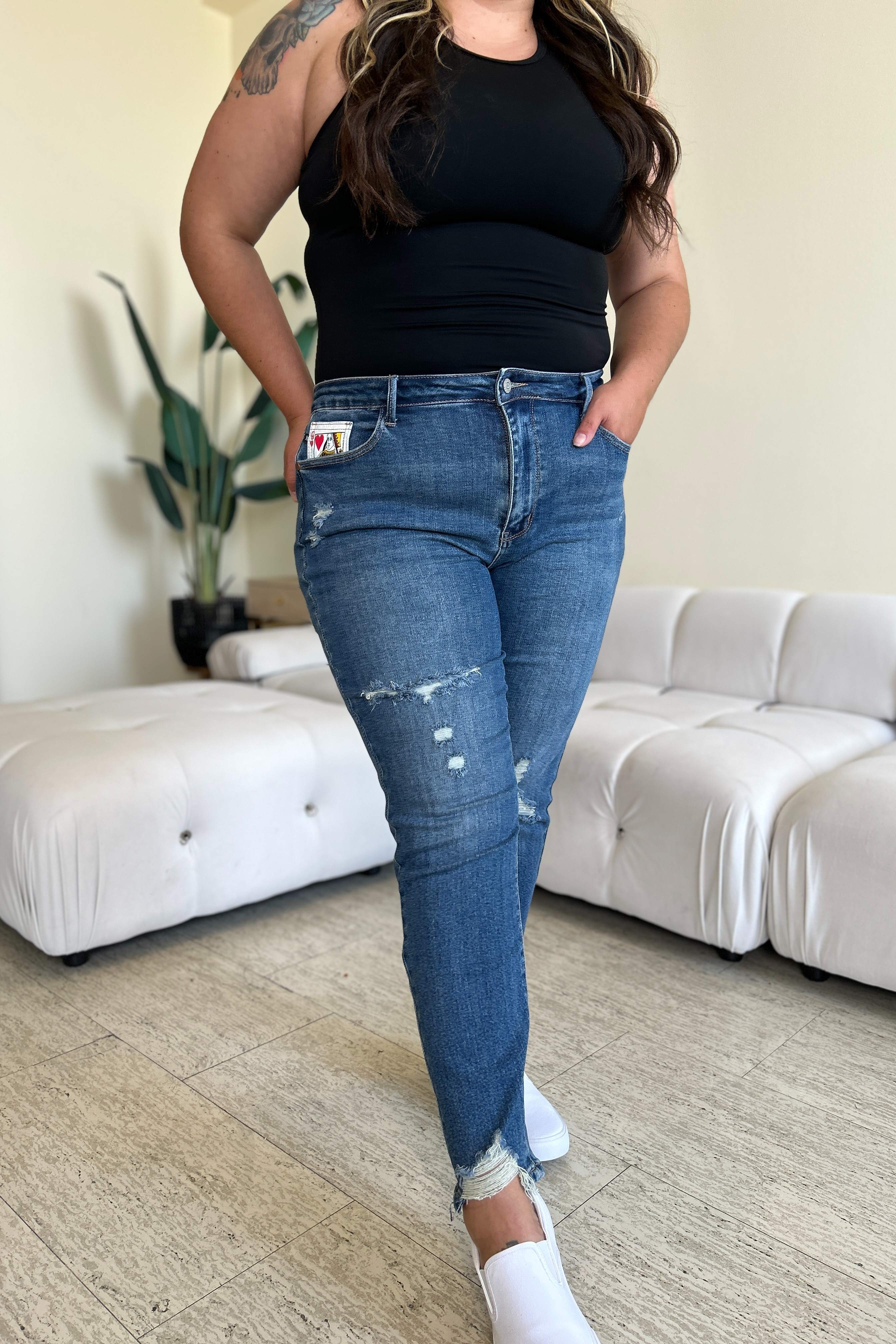 Two women stand side by side in a living room, both wearing stylish jeans and tank tops. One woman is pointing at the distinctive back pocket of her Judy Blue Full Size Queen Of Hearts Coin Pocket BF Jeans, while the other smiles.