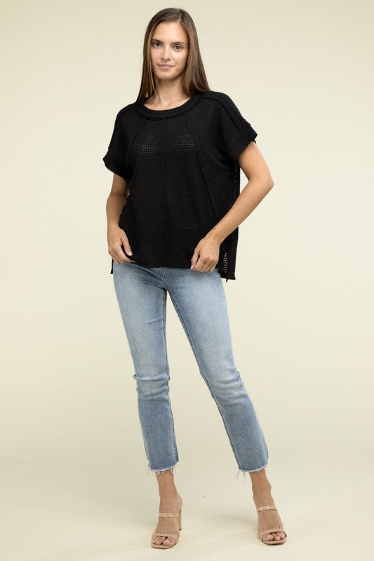 A woman with long brown hair is wearing a purple Brushed Waffle Exposed-Seam Short Sleeve Top and blue jeans, standing against a plain beige background. This casual wardrobe choice, with its short sleeves and side slits, is perfect for everyday wear.