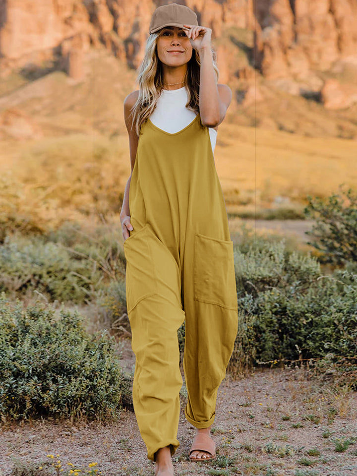 A woman wearing the Double Take Full Size Sleeveless V-Neck Pocketed Jumpsuit in coral and a beige cap is walking outdoors on a clear day, with a rocky landscape in the background.