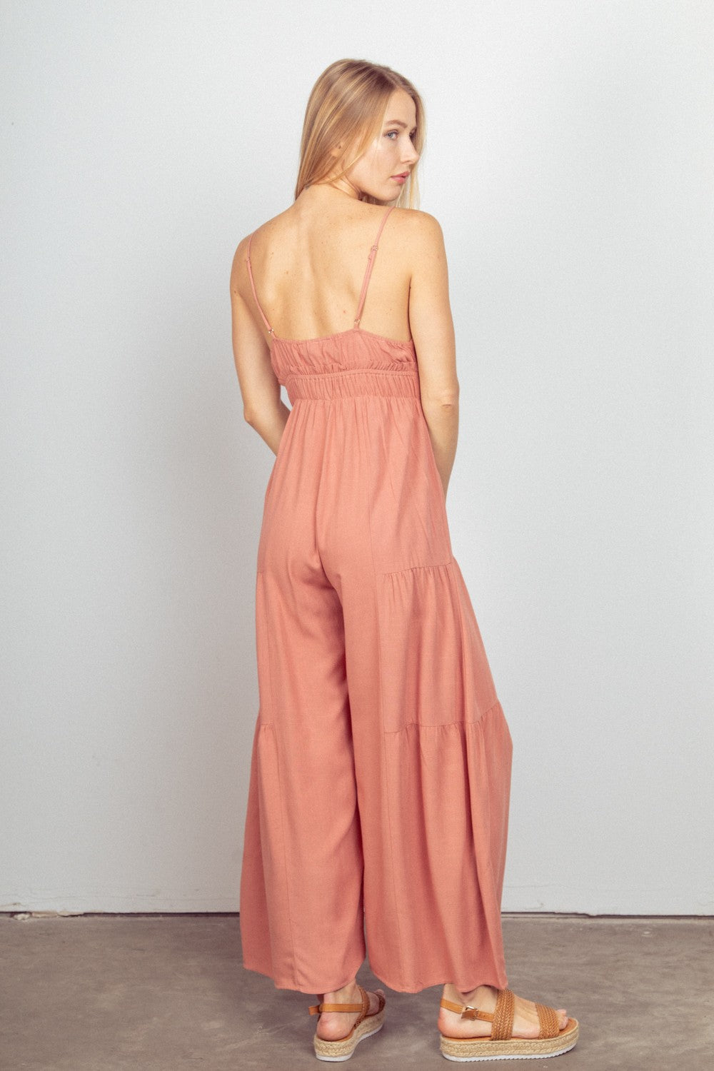 A woman stands against a plain background, wearing the VERY J Sleeveless Ruched Wide Leg Jumpsuit in pink, featuring an elastic waist and adjustable straps. She completes the look with crossover sandals.