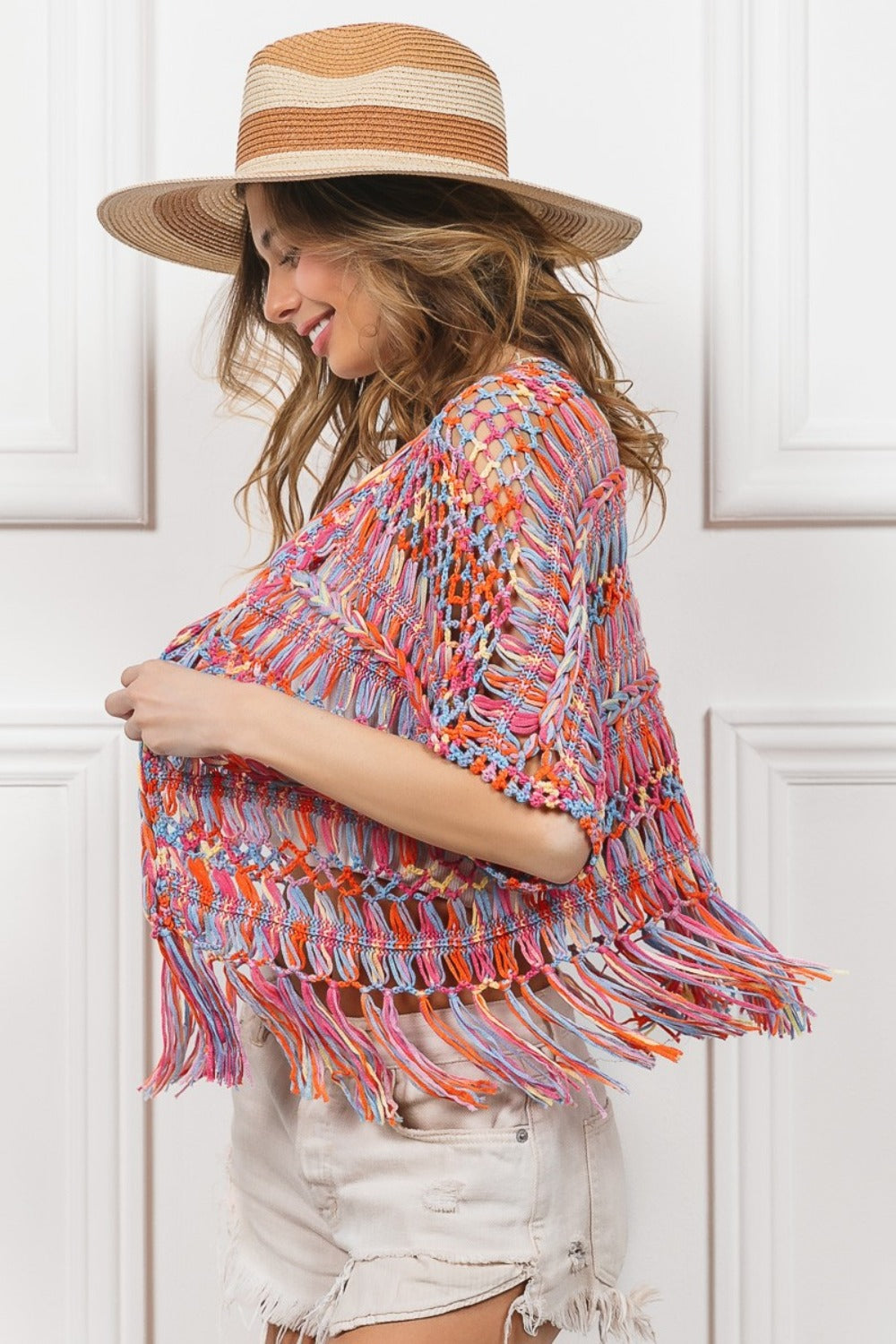 A woman wearing a BiBi Open Front Fringed Crop Knit Cardigan, a white bandeau, distressed white shorts, and a wide-brimmed straw hat stands against a white paneled background, smiling with one hand on her hat.