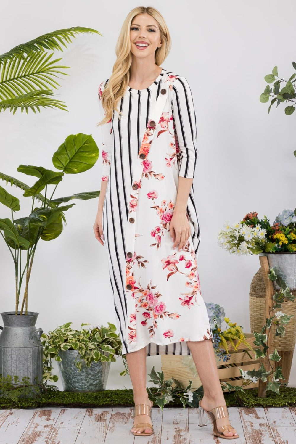 Dressed in the Celeste Full Size Floral Striped Contrast Midi-Dress with Pockets, a person stands elegantly among potted plants and flowers on a wooden floor, highlighting the dress's versatile styling options.