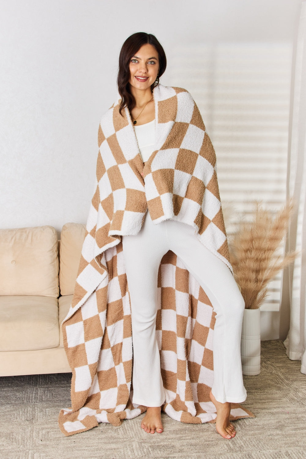 A person stands holding a large Cuddley Checkered Decorative Throw Blanket, smiling, in a room with a sofa and a potted plant. The blanket features a black and white checkerboard pattern, is made from 100% polyester, and imported for quality.