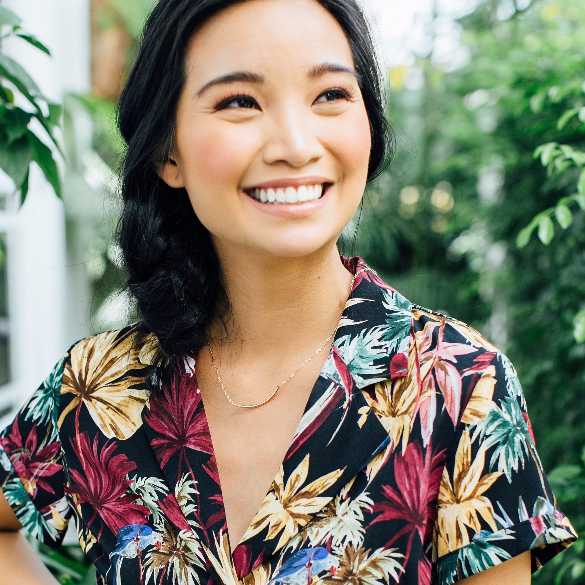 Amidst lush greenery, a person smiles outdoors, wearing a floral shirt and the delicate Talitha Necklace in 14k gold fill.