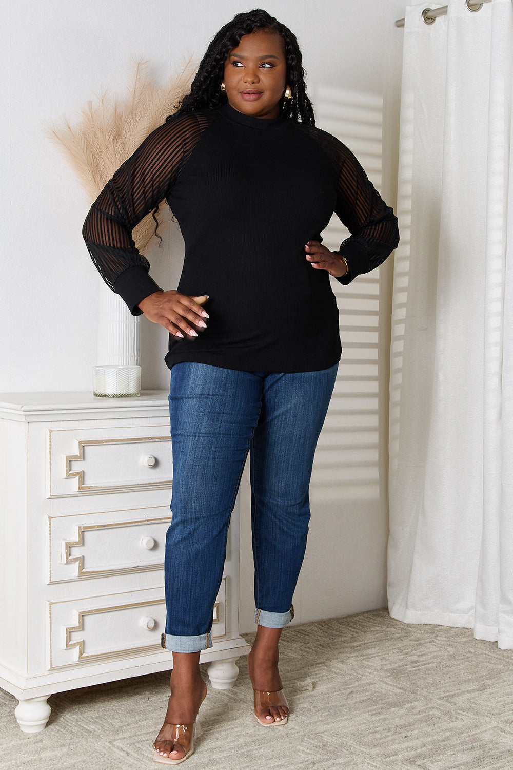 A woman with long dark hair stands indoors wearing a versatile wardrobe piece: the Double Take Round Neck Raglan Sleeve Blouse paired with blue jeans, her right hand in her pocket. A white vase with pampas grass is on the table next to her.