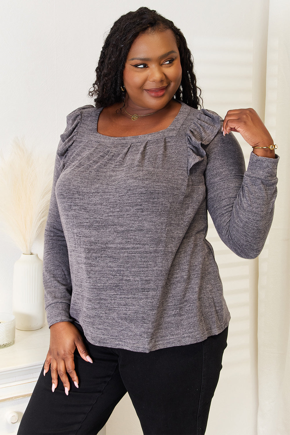 A woman wearing a Double Take Square Neck Ruffle Shoulder Long Sleeve T-Shirt in pink and blue jeans stands with one hand in her pocket, smiling. She has long curly hair and wears a necklace and bracelet. She stands in a room with white decor.