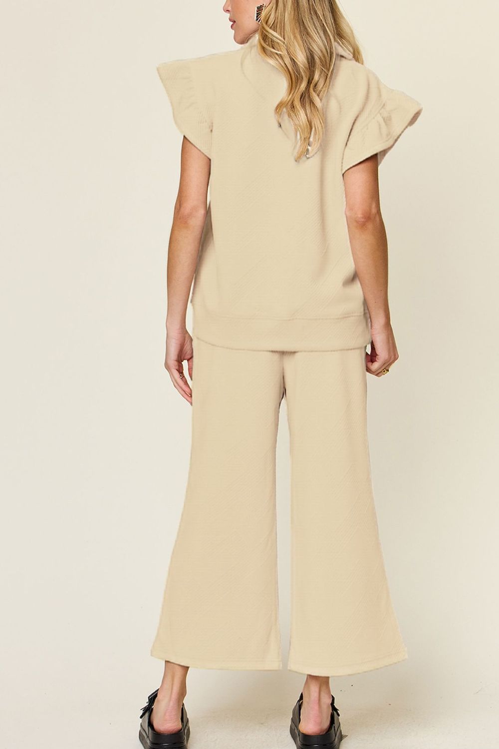 A woman stands wearing the Double Take Texture Ruffle Short Sleeve Top and Drawstring Wide Leg Pants Set in a slightly stretchy, light gray fabric with zipper detail. The two-piece outfit is complemented by black sandals, and she has one hand in her pocket. Machine wash cold for easy care.