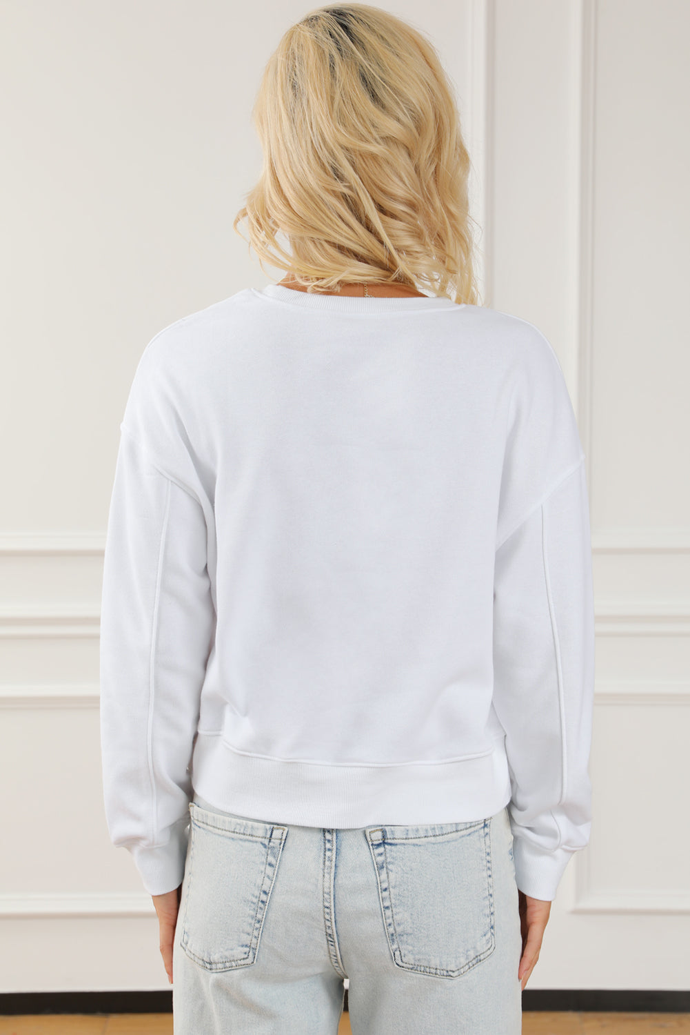 A person with long blonde hair is standing with their back to the camera, wearing a White BEACH BABE Slogan Graphic Casual Sweatshirt and light blue jeans.