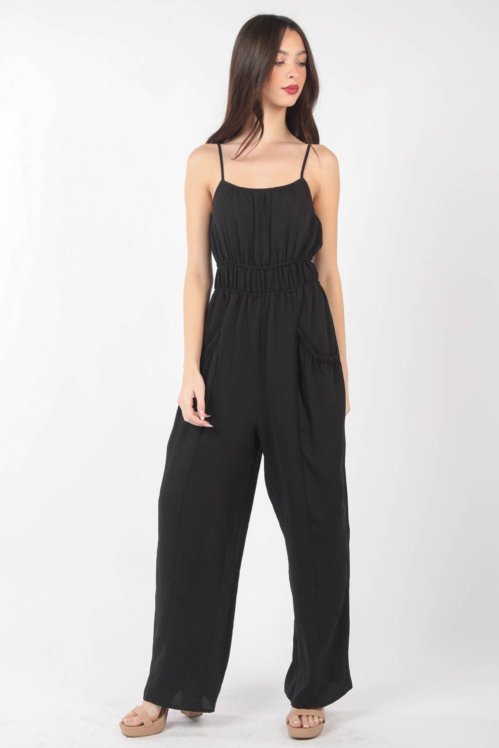 A woman stands against a plain backdrop wearing the VERY J Pintuck Detail Woven Sleeveless Jumpsuit. The black jumpsuit has thin straps, wide-leg pants, and is crafted from woven fabric with subtle pintuck details. She has her hands in her pockets and is looking at the camera.