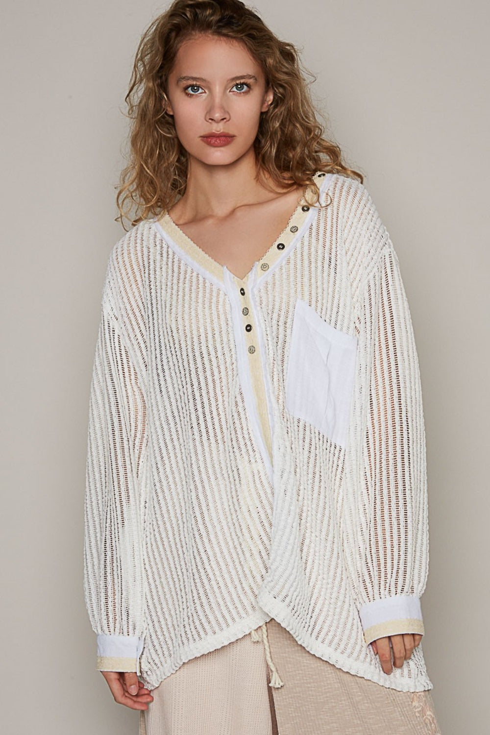 A person with wavy hair wearing a loose, bohemian-style POL V-Neck Long Sleeve Crochet Top in a sheer, striped white design paired with beige pants stands against a plain background.