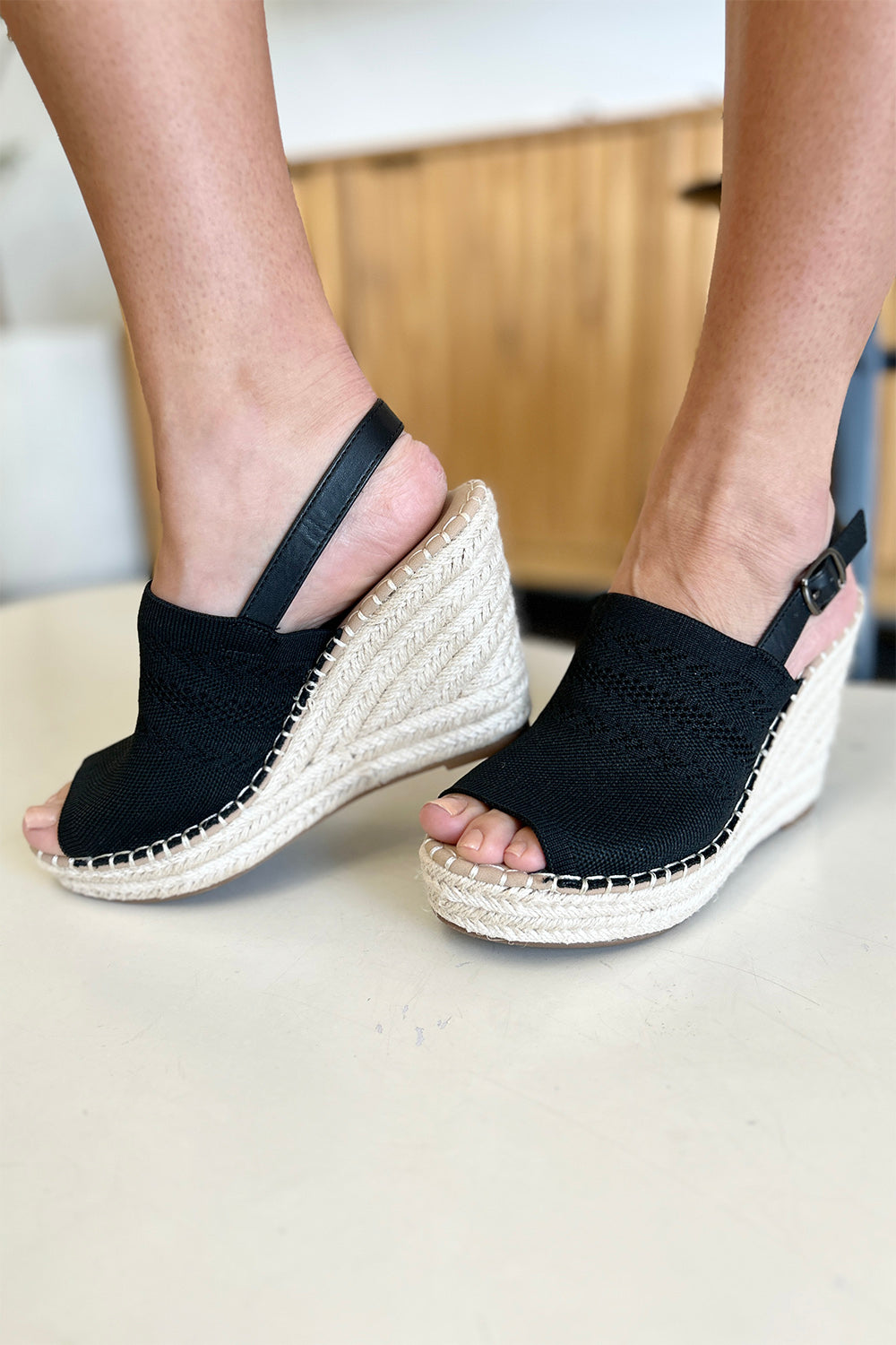 Close-up of a person's feet wearing chic black Forever Link Peep Toe High Heel Sandals with a braided espadrille base and adjustable heel straps. The background is blurred, making these sandals the stylish choice for any outfit.