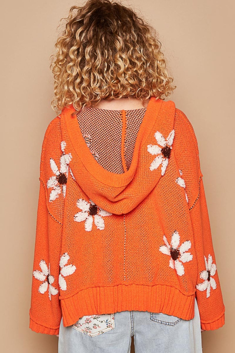 A person with curly blond hair is wearing the POL Floral Pattern Hooded High-Low Sweater in orange, featuring white and brown flower patterns against a solid beige background, paired with light blue jeans.