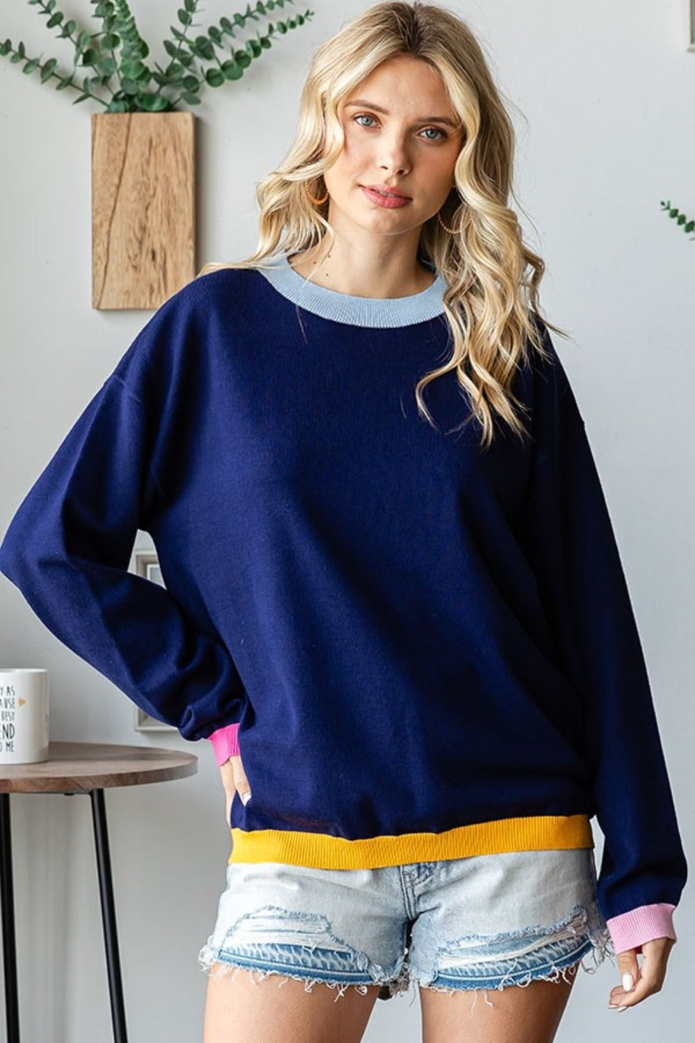 A person with long blonde hair wearing a First Love Contrast Ribbed Round Neck Long Sleeve Sweater and light denim shorts stands indoors near a small table and plant.