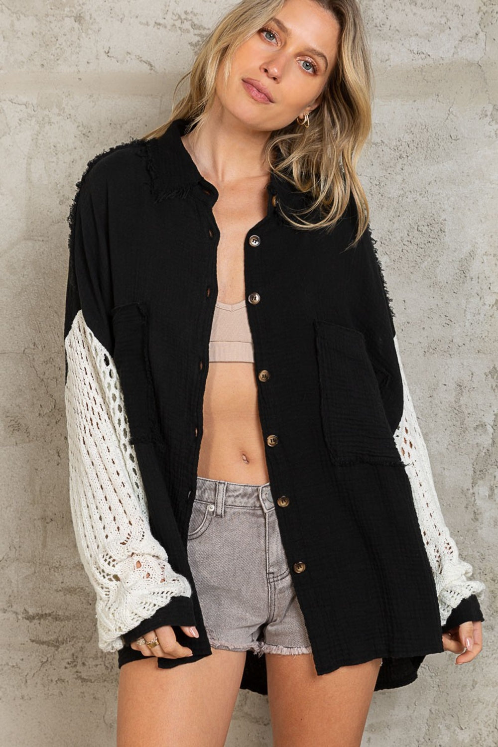 A woman with blonde hair stands against a gray textured wall, wearing the POL Openwork Lantern Sleeve Button Down Gauze Shirt—an elegant, lightweight, and breathable black button-up shirt with white knit sleeves. She pairs it with a beige top and gray shorts, creating a chic ensemble.