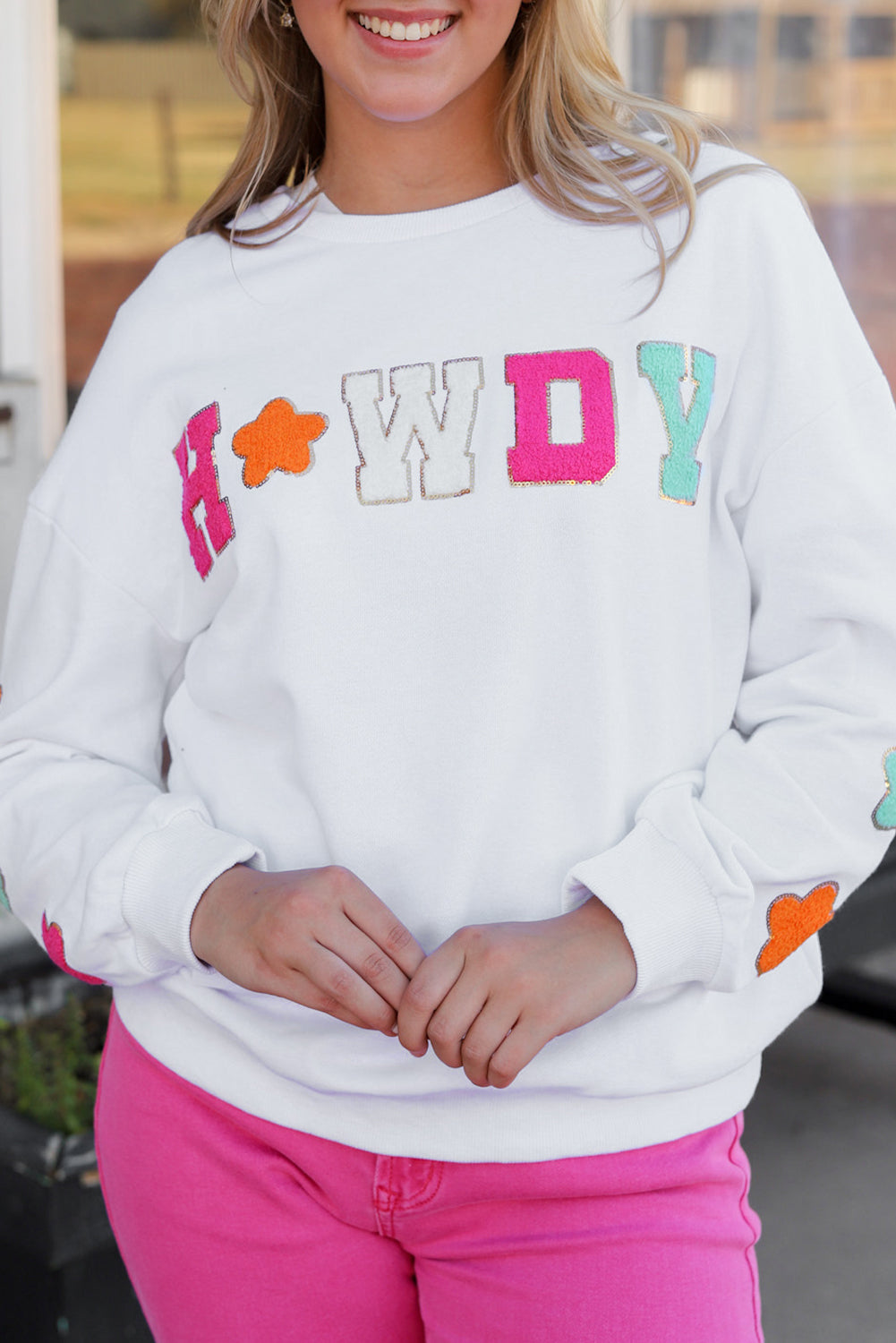 A person poses in the White Howdy Glitter Chenille Patch Graphic Casual Sweatshirt, adorned with colorful star patterns and the letters "WD." She wears light denim shorts and has shoulder-length blonde hair.