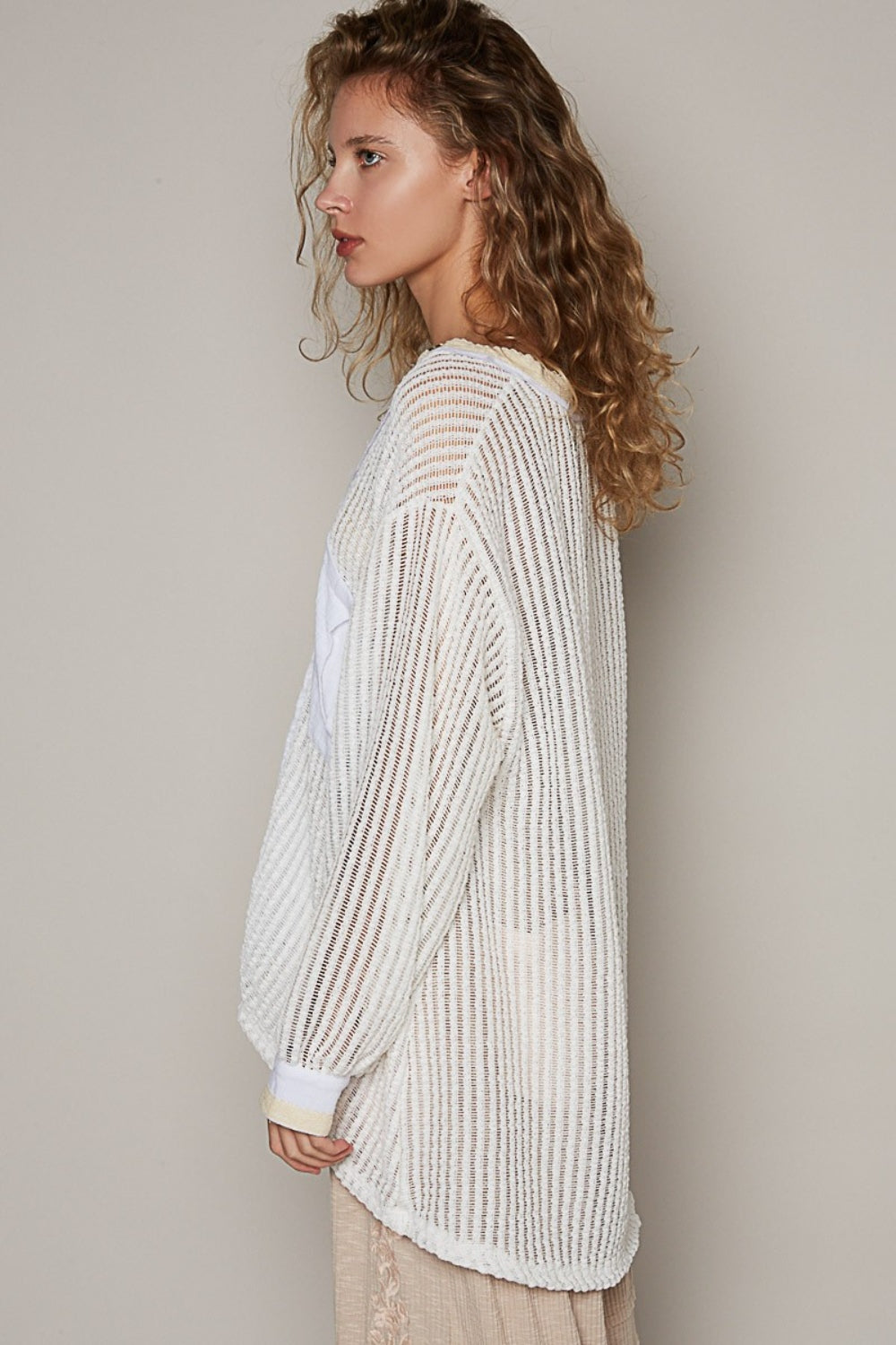 A person with wavy hair wearing a loose, bohemian-style POL V-Neck Long Sleeve Crochet Top in a sheer, striped white design paired with beige pants stands against a plain background.