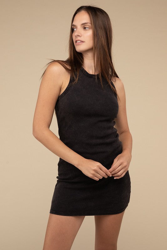 A woman is seated, wearing the Acid Washed Ribbed Mini Dress in a faded red hue, along with a necklace. Her right arm rests on a white surface as her straight hair frames her neutral expression.