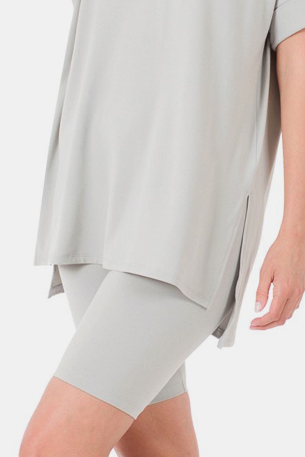 A woman is wearing the Zenana Full Size V-Neck Short Sleeve Slit T-Shirt and Shorts Set in a light gray color. The loose-fitting, highly stretchy outfit features a V-neck t-shirt with short sleeves and matching knee-length shorts. She is smiling with her hand raised near her head.