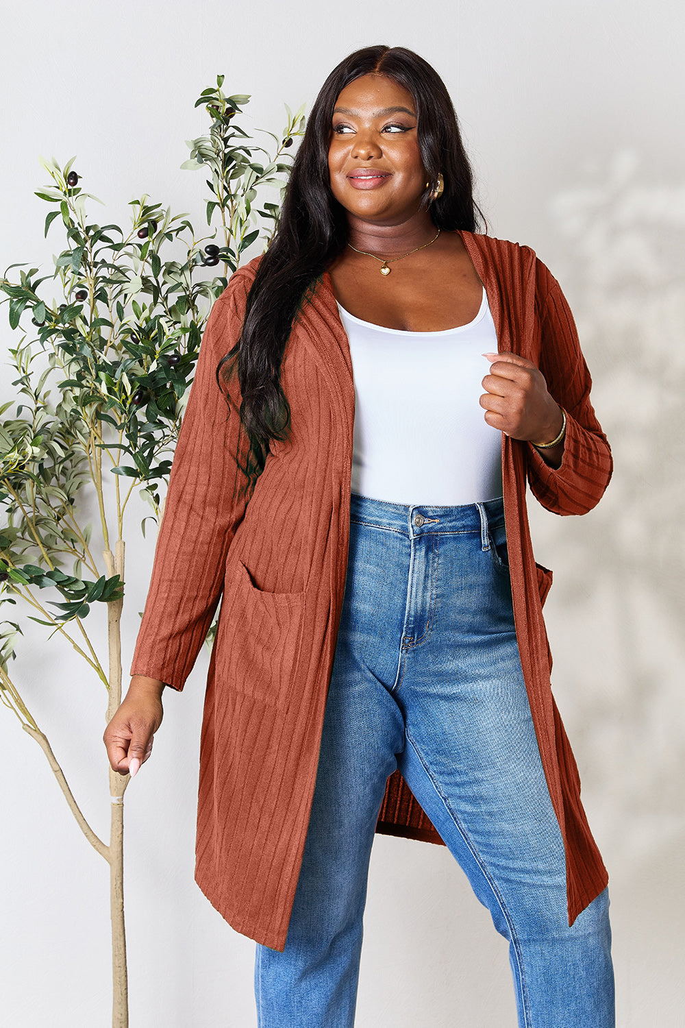 A woman wearing the Basic Bae Full Size Hooded Sweater Cardigan in black, paired with a white top and khaki pants, stands in front of a light background accented by a leafy plant on her left. With one hand tucked into her pocket, she gazes to her right.