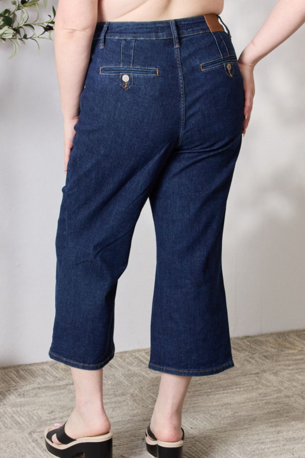 A person wearing the Judy Blue Full Size High Waist Cropped Wide Leg Jeans, which are renowned for their tummy control design, and nude block heel sandals stands near a potted plant.