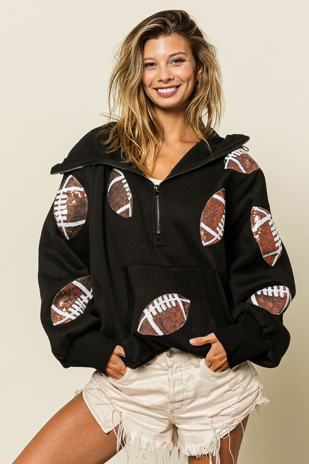 A person wearing a BiBi Sequin Football Half Zip Hoodie and beige shorts stands smiling in front of a neutral background with dried plants.