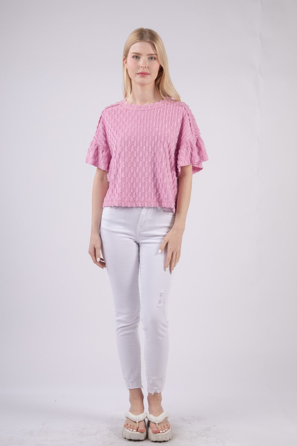 Person wearing the VERY J Full Size Texture Ruffle Short Sleeve Top, a chic and feminine textured pink blouse, paired with white pants, standing against a plain background.