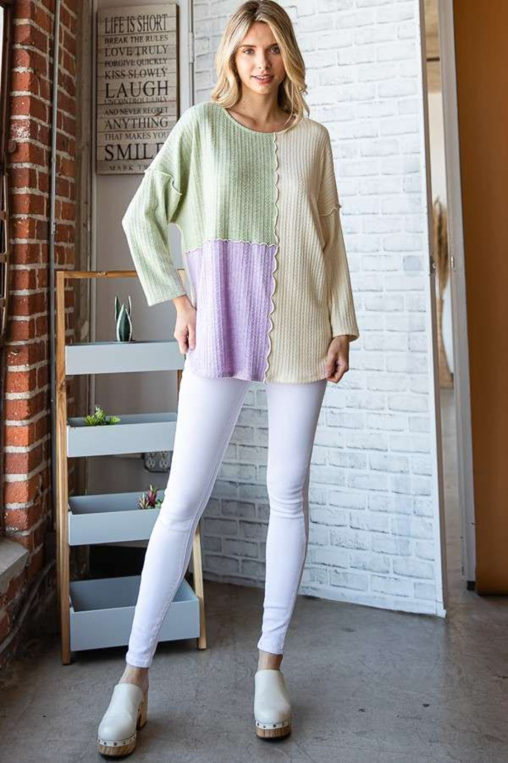 A person dressed in the Culture Code Color Block Round Neck Dropped Shoulder Blouse, featuring pastel shades of green, ivory, and lavender, stands in a room with a decorative sign on the brick wall.