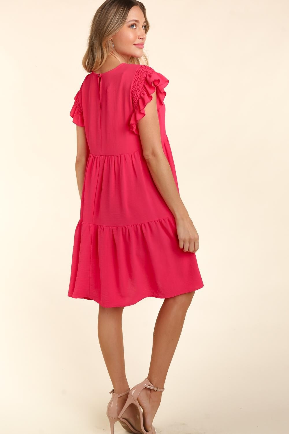 A woman with long, wavy hair wearing a bright pink Haptics Full Size Smocking Ruffle Short Sleeve Dress with Pockets, featuring ruffled sleeves and a smocked waist, stands smiling with arms crossed in front of a neutral background.