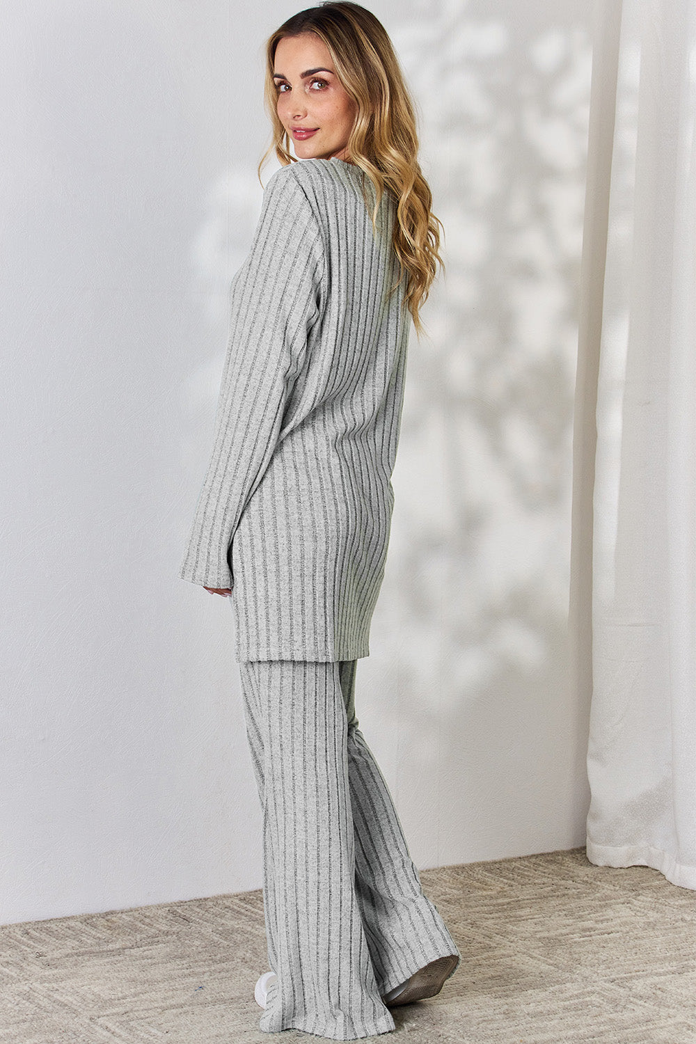 A person wearing the Basic Bae Full Size Ribbed High-Low Top and Wide Leg Pants Set in light gray, made from stretchy material, stands indoors in front of a white curtain.